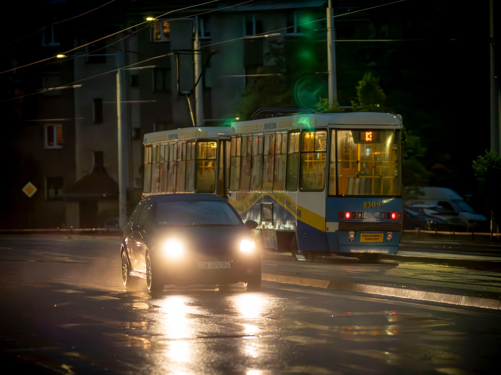 A car and tram pass through a rain-soaked street in Wrocław, Poland at night, showcasing urban transit.
