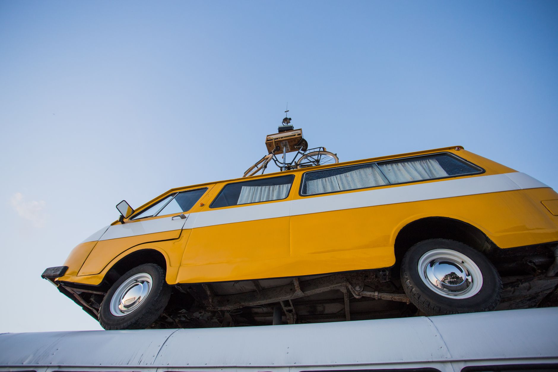 A yellow vintage van mounted on a rooftop against a clear blue sky captured from a unique angle.