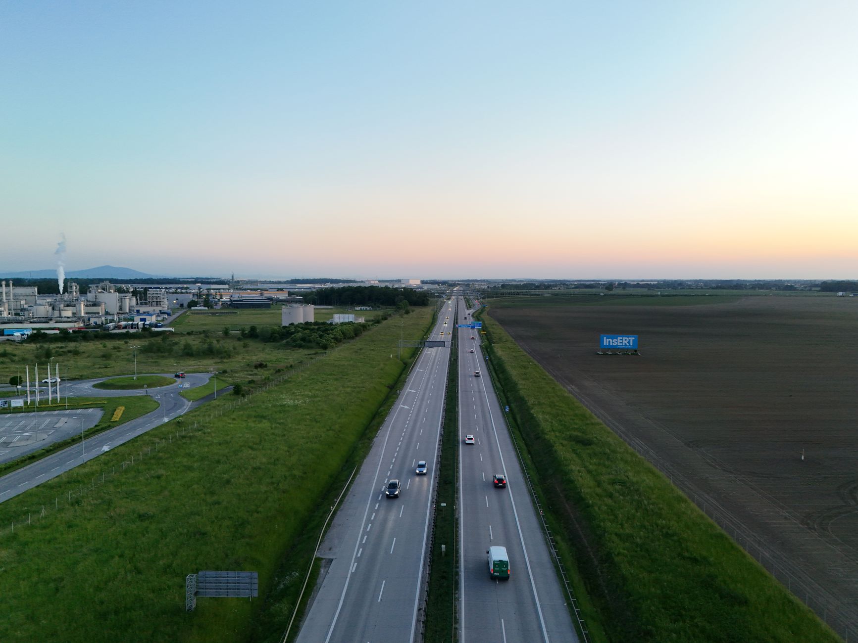 Aerial photograph shows a highway at sunset near a factory and fields.