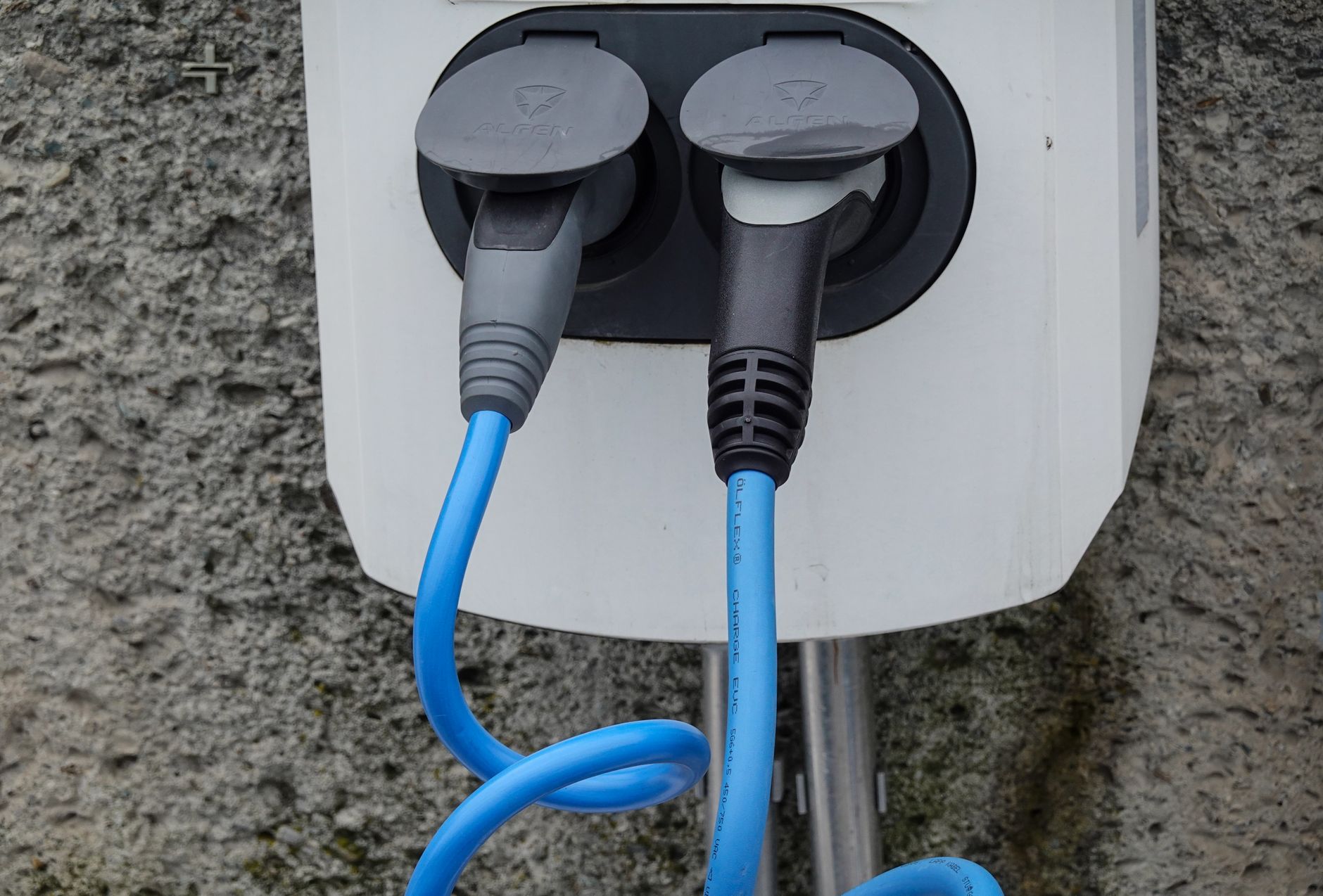Close-up of an electric vehicle charging station with two cables in sockets against a textured wall.