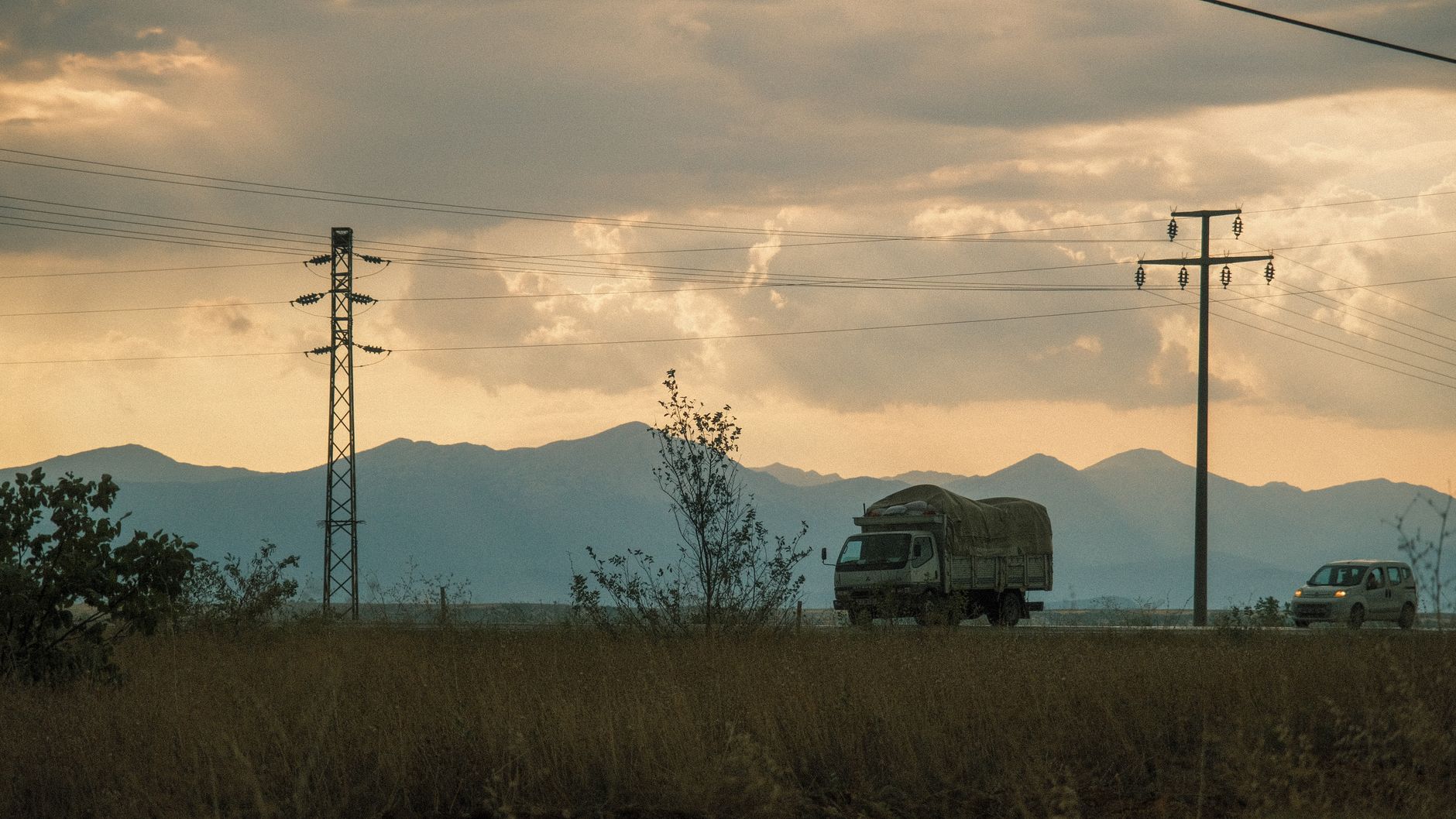 Trucks travel on a rural road at sunset, framed by power lines and distant mountains.