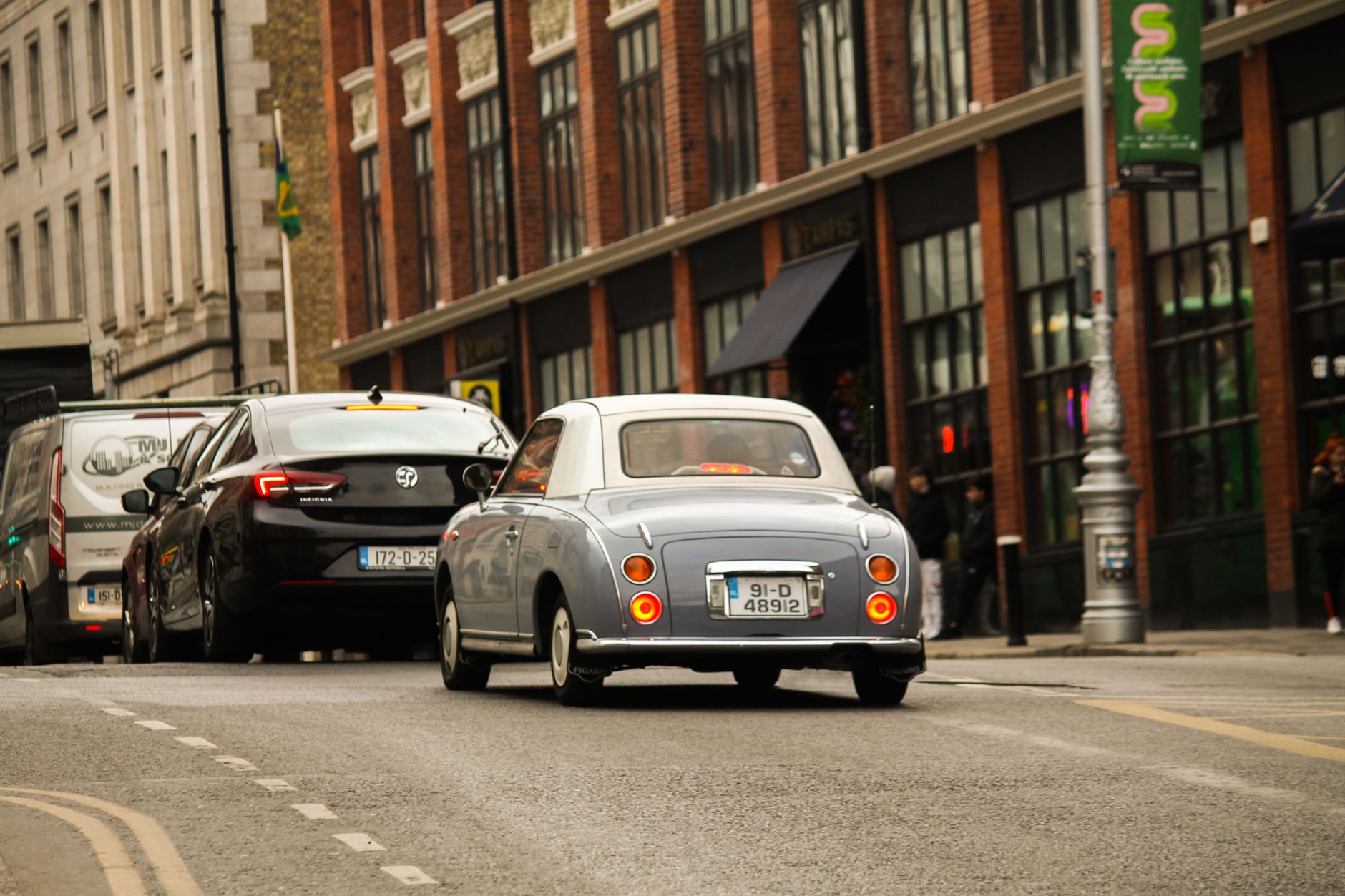 A classic Nissan Figaro driving through a busy urban street lined with brick buildings.