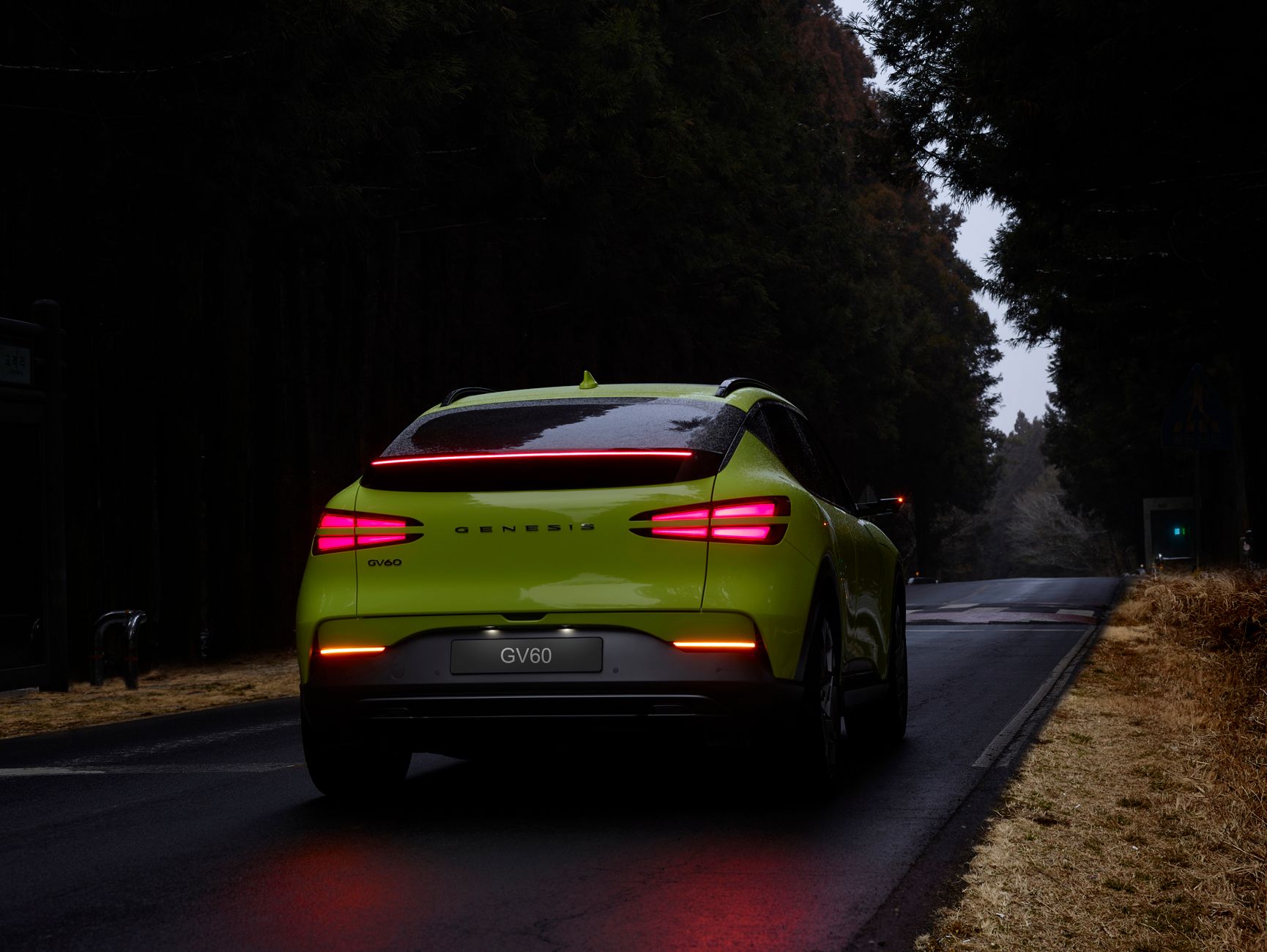 Lime green Genesis GV60 SUV traveling on a dark forest road, captured from behind.